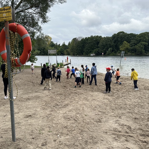 Gruppenspielende Kinder am Sandstrand eines Sees, mit einem Rettungsring im Vordergrund und Bootssteg im Hintergrund.