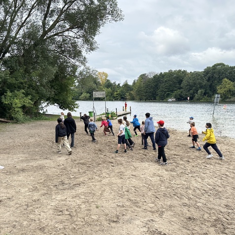 Gruppe von Kindern spielt am Ufer eines Gew&auml;ssers, w&auml;hrend eine Betreuerin in der N&auml;he steht.