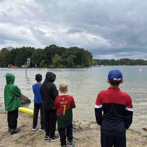 F&uuml;nf Kinder stehen am Ufer eines Gew&auml;ssers und beobachten ein Boot unter grauem Himmel.