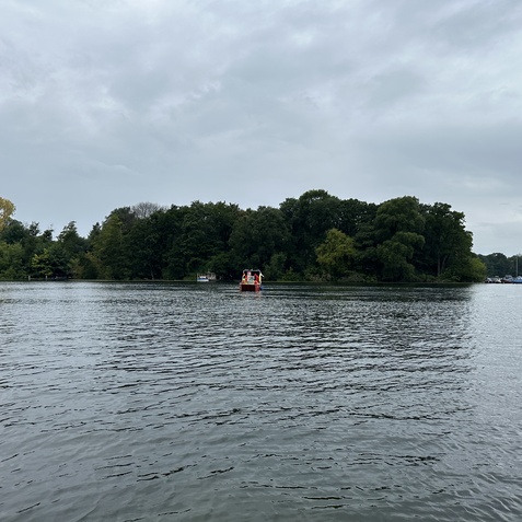 Ein Boot f&auml;hrt auf einem ruhigen Gew&auml;sser, umgeben von gr&uuml;nen Ufern und bewaldeten Bereichen unter einem grauen Himmel.
