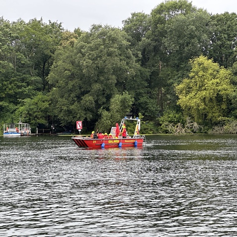 Rettungsboot mit zwei Personen auf einem Gew&auml;sser, umgeben von gr&uuml;ner Ufervegetation und einem weiteren Boot im Hintergrund.