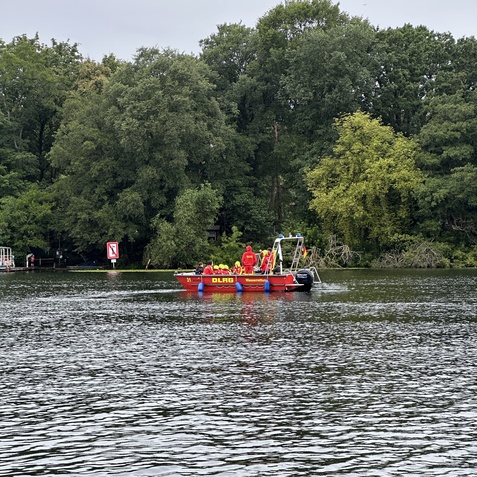 Rettungsboot mit DLRG-Logo, besetzt von mehreren Personen, f&auml;hrt auf einem Wasserweg zwischen gr&uuml;nen B&auml;umen.