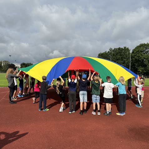 Gruppenspiel von Kindern, die einen bunten Fallschirm auf einem Sportplatz gemeinsam halten und bewegen.