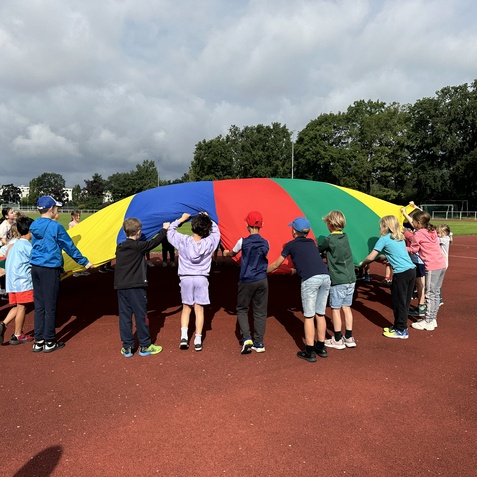 Kinder halten eine bunte Gruppenspielplane auf einem roten Sportplatz, umgeben von B&auml;umen und Himmel.