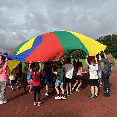 Gruppenspiel von Kindern, die gemeinsam einen bunten Fallschirm auf einem Sportplatz halten.
