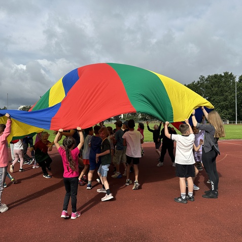 Kinder halten ein gro&szlig;es, bunten Fallschirm auf einem Sportplatz und spielen im Freien unter bew&ouml;lktem Himmel.