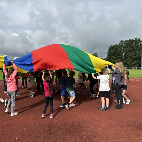 Kinder und Jugendliche spielen mit einem bunten Fallschirm auf einem Sportplatz unter wolkigem Himmel.