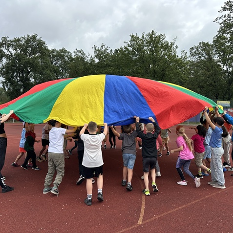 Gruppe von Kindern und Erwachsenen hebt ein buntes Spielzelt auf einem roten Sportplatz unter bew&ouml;lktem Himmel.