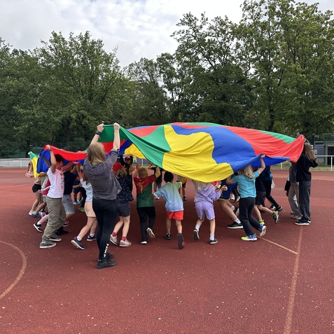 Gruppe von Kindern spielt mit einem bunten Fallschirm auf einem Sportplatz im Freien. B&auml;ume im Hintergrund.