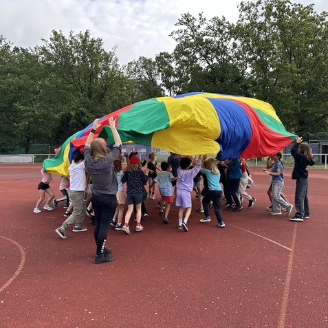 Kinder spielen mit einem bunten Fallschirm auf einem Sportplatz, umgeben von B&auml;umen und einem grauen Himmel.