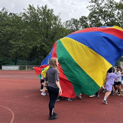 Kinder spielen mit einem gro&szlig;en, bunten Fallschirm auf einem Sportplatz unter bew&ouml;lktem Himmel.