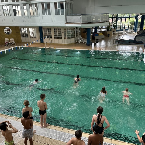 Gruppe von Kindern schwimmt in einem Hallenbad, w&auml;hrend andere am Beckenrand zuschauen.