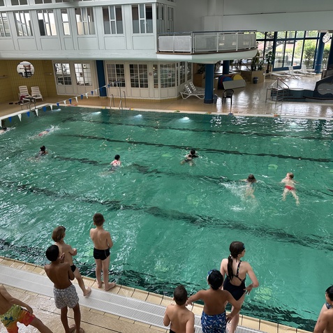 Kinder schwimmen im Indoor-Pool, w&auml;hrend einige am Beckenrand stehen und zuschauen. Klarer Wasser- und Raumkontrast.