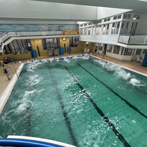 In einem Schwimmbad spritzen mehrere Schwimmer im Wasser, umgeben von einer hellen Umgebung mit Fensterfront.