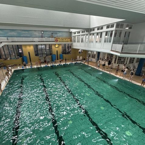 Gruppen von Schwimmern stehen am Rand eines Innenpools, Wasser spiegelt den Raum in hellgr&uuml;nen T&ouml;nen wider.