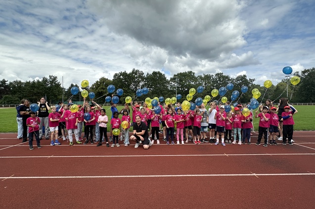 Gruppe von Kindern in pinken T-Shirts mit bunten Ballons auf einer Laufbahn, unter wolkigem Himmel.