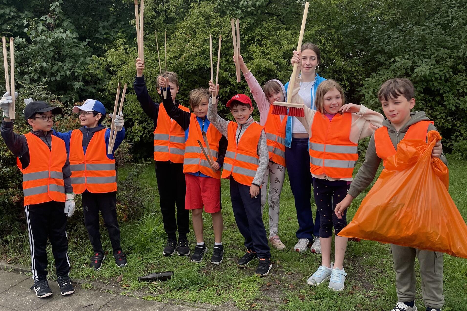 Gruppe von Kindern in orangefarbenen Warnwesten mit M&uuml;llzangen und einem M&uuml;llsack, stehend in einer Gr&uuml;nanlage.