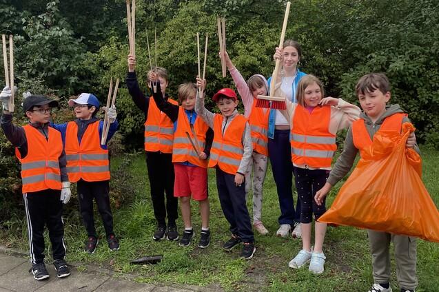 Gruppe von Kindern in orangefarbenen Warnwesten mit Müllzangen und einem Müllsack, stehend in einer Grünanlage.