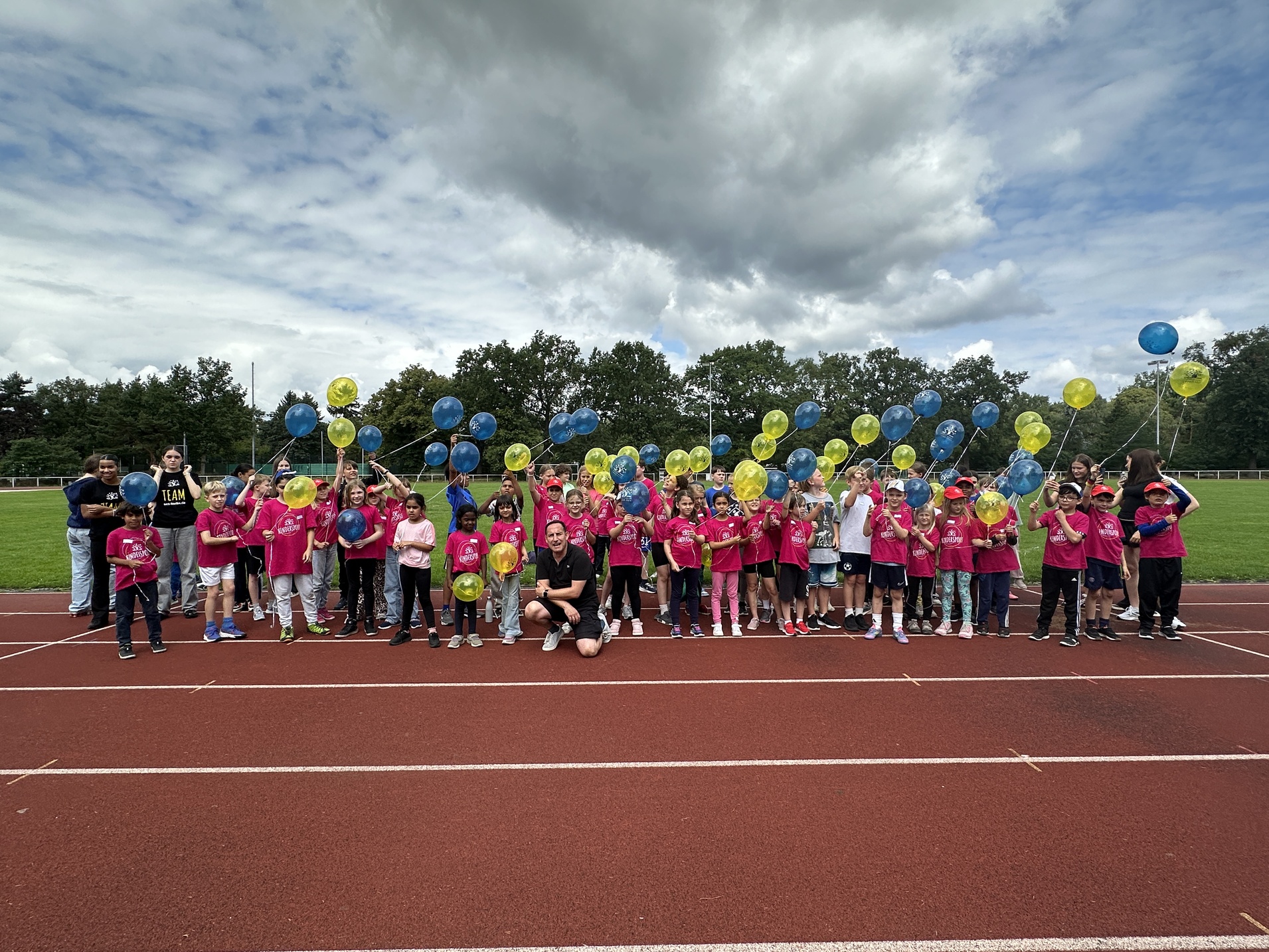 Gruppe von Kindern in pinken T-Shirts mit bunten Ballons auf einer Laufbahn, unter wolkigem Himmel.