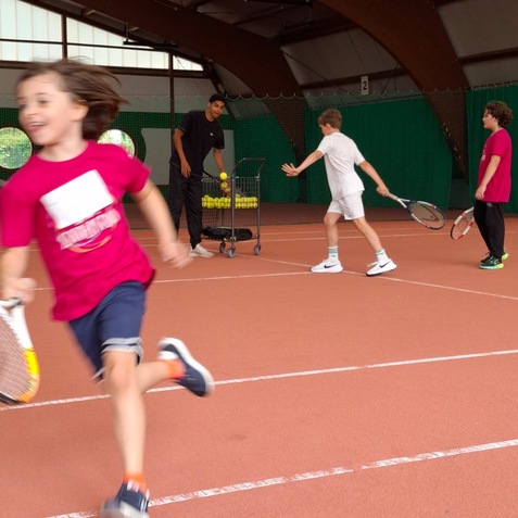Kinder spielen Tennis in einer Halle, w&auml;hrend ein Trainer B&auml;lle in einem Wagen vorbereitet.