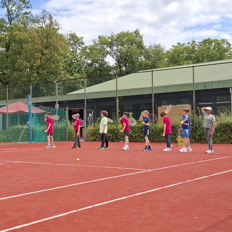 Gruppe von Kindern steht auf einem Tennisplatz in einer Reihe, jeder h&auml;lt einen Tennisschl&auml;ger in der Hand.