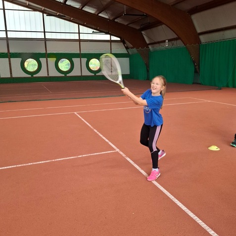 M&auml;dchen in blauer Sportkleidung schl&auml;gt mit einem Tennisschl&auml;ger einen Ball in einer Tennishalle auf rotem Belag.