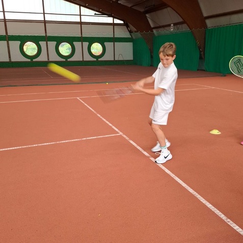Junge in wei&szlig;em Tennisoutfit schl&auml;gt einen Tennisball auf einem Indoor-Tennisplatz mit ockerfarbenem Boden.