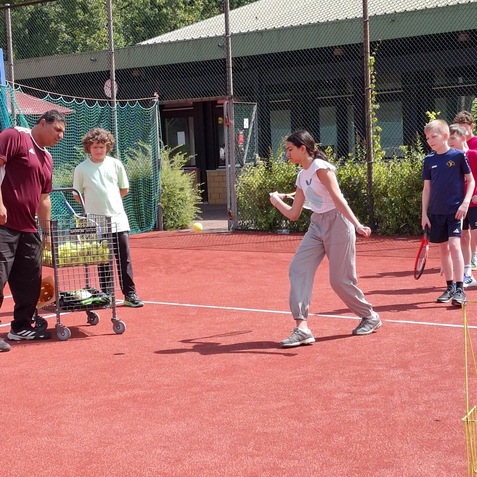 Gruppe von Kindern und einem Trainer auf einem Tennisplatz; ein M&auml;dchen schl&auml;gt einen Ball mit einem Schl&auml;ger.