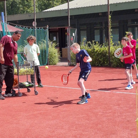 Vier Kinder trainieren Tennis auf einem Au&szlig;entennisplatz, ein Trainer steht mit einem Ballwagen daneben.