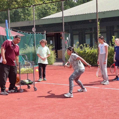 Tennisunterricht: Trainer mit Kindern auf einem roten Platz, ein M&auml;dchen &uuml;bt mit Schl&auml;ger. Hintergrund mit Netzen und B&auml;umen.