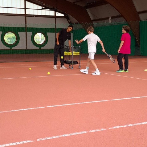 Trainer zeigt zwei Kindern beim Tennisunterricht, ein Kind h&auml;lt einen Schl&auml;ger, der Ballkorb ist im Hintergrund sichtbar.