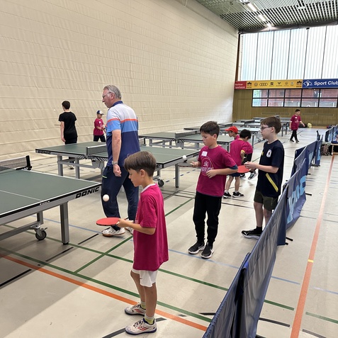 Gruppe von Kindern in pinken T-Shirts &uuml;bt Tischtennis unter Anleitung eines Trainers in einer Sporthalle.