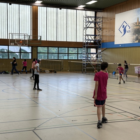Gruppentraining in einer Sporthalle mit Kindern, die Badminton spielen, w&auml;hrend andere auf der Bank sitzen.