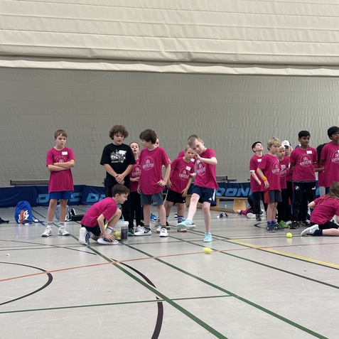 Gruppe von Kindern in pinken T-Shirts bei einer Sportveranstaltung in einer Halle, einige spielen mit einem Tennisball.