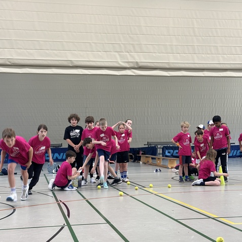 Gruppe von Kindern in pinken T-Shirts trainiert in einer Sporthalle, mehrere Tennisb&auml;lle liegen auf dem Boden.