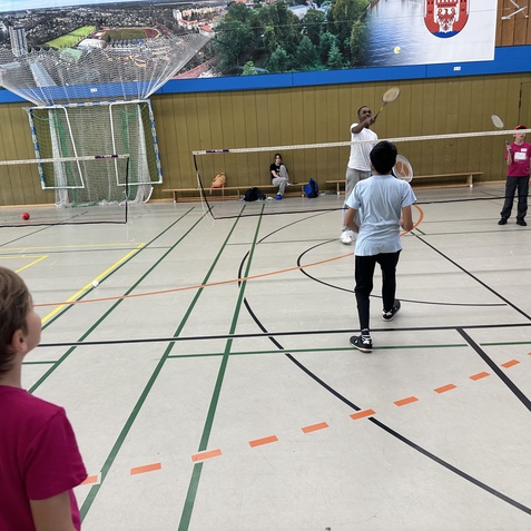 Gruppen von Kindern spielen Badminton in einer Sporthalle, w&auml;hrend ein Trainer einem Jungen hilft, einen Aufschlag zu &uuml;ben.