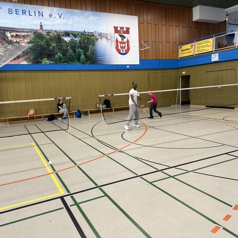 Kinder und Erwachsene spielen Badminton in einer Sporthalle mit Netzen und Banden. Im Hintergrund ist ein Wandbanner sichtbar.