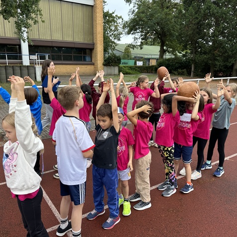 Gruppe von Kindern in pinken T-Shirts mit erhobenen H&auml;nden, einige halten Basketballs, auf einem Sportplatz.