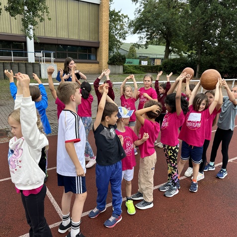 Gruppe von Kindern in pinken T-Shirts, die auf einem Sportplatz Basketball spielen und ihre H&auml;nde heben.
