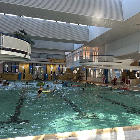 Innenansicht eines Schwimmbads mit schwimmenden Menschen, Liegen und einem zweiten Stock mit Fenstern.