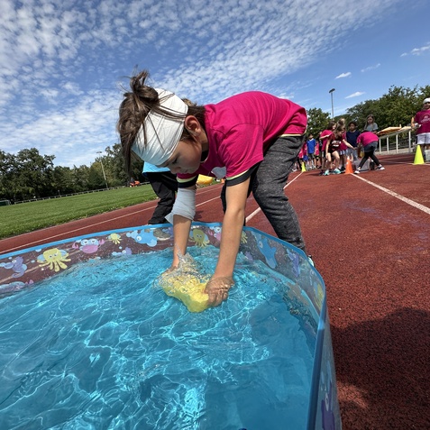 Kind mit einem Bartuch greift nach einem gelben Schwamm im blauen Wasser eines Planschbeckens auf einem Sportplatz.