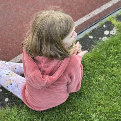 Mädchen sitzt auf dem Gras am Rand einer Laufbahn, trägt einen rosa Hoodie und Schlafanzughosen mit Entenmotiven.