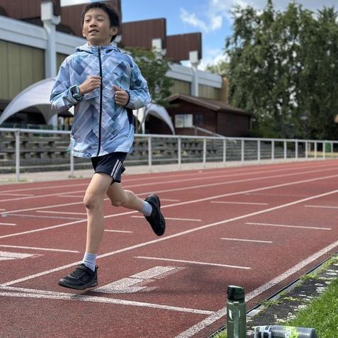 Junge läuft auf einer Leichtathletikbahn, Wasserflaschen liegen am Rand, im Hintergrund Gebäude und Bäume.