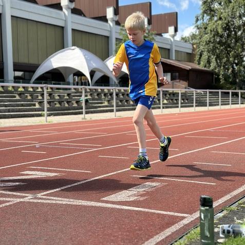 Ein Junge in blau-gelbem Sportanzug läuft auf einer Leichtathletikbahn, im Hintergrund eine Sporthalle.
