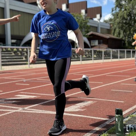 Ein Mädchen mit Brille läuft auf einer roten Tartanbahn, trägt ein blaues T-Shirt und schwarze Leggings.