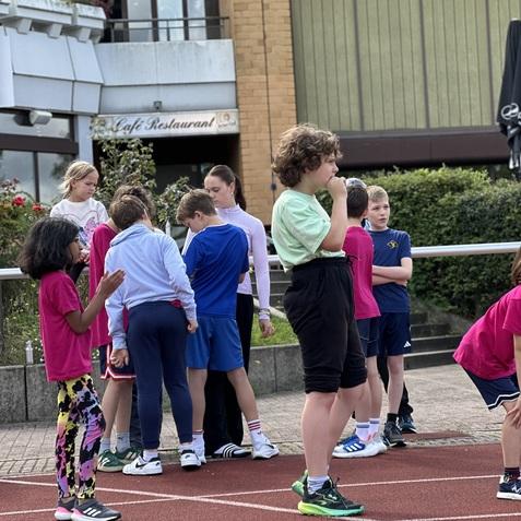 Gruppe von Kindern in Sportkleidung auf einer Laufbahn, einige stehend, andere im Gespräch. Hintergründe sichtbar.