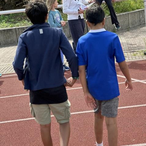Gruppe von Kindern auf einer Laufbahn; zwei Jungen in Sportkleidung gehen, während andere Mädchen aktiv sind.
