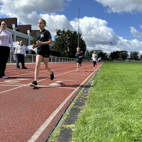 Schülerinnen laufen auf einer Leichtathletikbahn, im Hintergrund stehen Zuschauer und Bäume unter einem blauen Himmel.