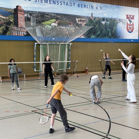 Gruppe von Kindern spielt Badminton in einer Sporthalle, mit Netzen und einem Wandbild im Hintergrund.