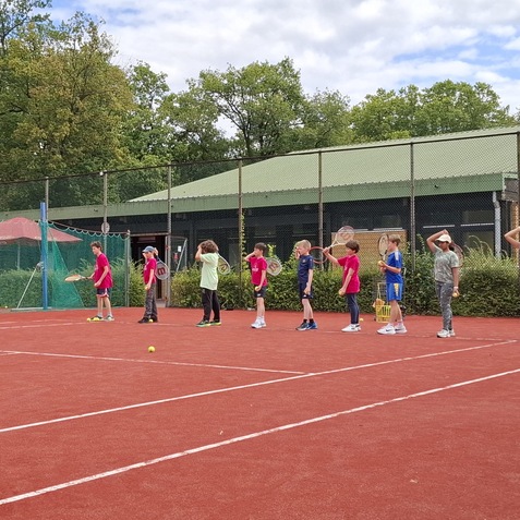 Gruppe von Kindern in Sportkleidung auf einem roten Tennisplatz, bereit zum Tennisunterricht, einige mit Schl&auml;gern in der Hand.